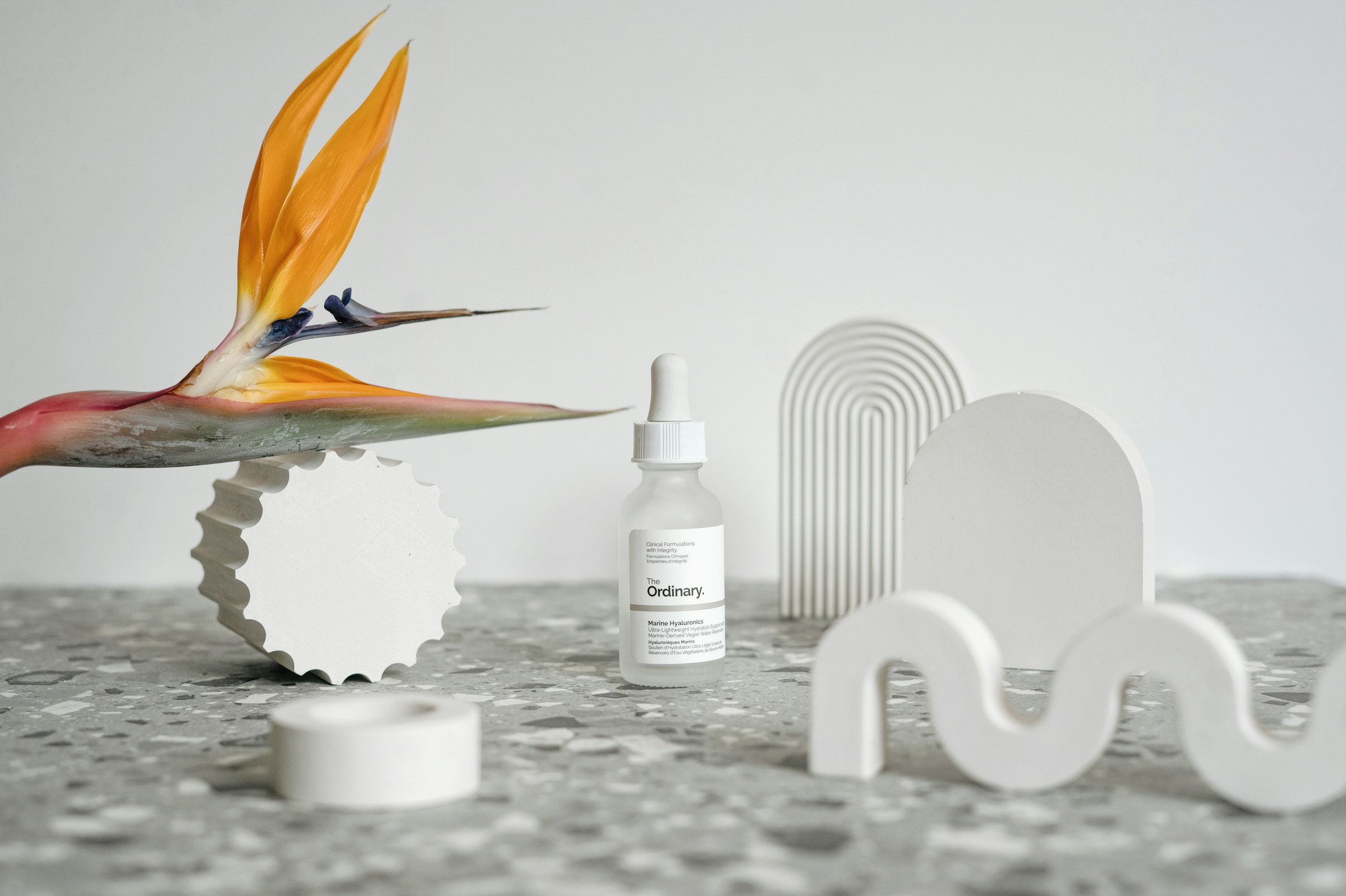 Ενυδατικός ορός προσώπου The Ordinary με σταγονόμετρο σε μίνιμαλ λευκό σκηνικό δίπλα σε άνθος στρελίτσιας, clean beauty και φυσικά καλλυντικά
